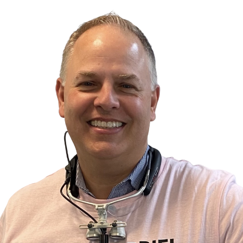 Smiling man wearing a pink polo with a head-mounted loupe and headset around his neck, looking at the camera.